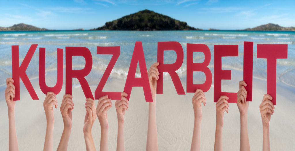 Hände halten Buchstaben "Kurzarbeit" vor Urlaubskulisse hoch