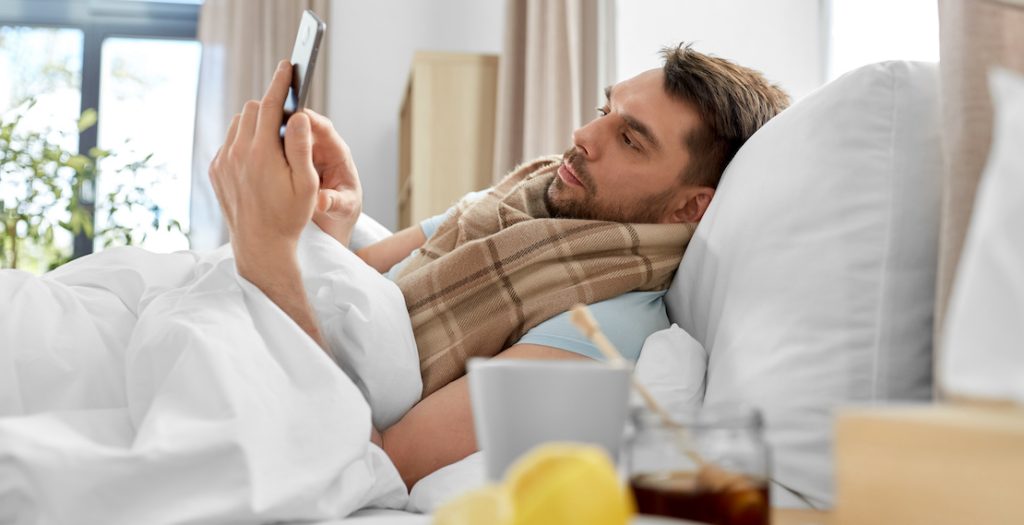 Mann liegt krank im Bett mit Smartphone.