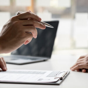 Hand hält Stift über einem Lebenslauf; gegenüber liegen die Hände eines Bewerbers auf dem Tisch.